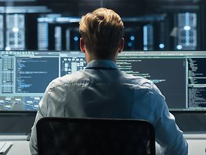 Mann sitzt vor mehreren PC-Bildschirmen, dunkler Hintergrund